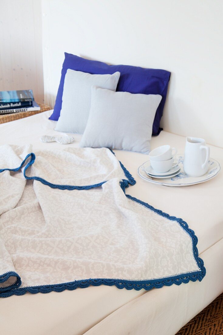 Decke mit blauer Häkelborte auf dem Bett mit Frühstücksgeschirr