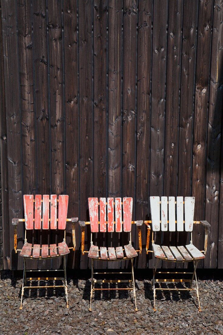 Three weathered chairs against wooden wall outdoors