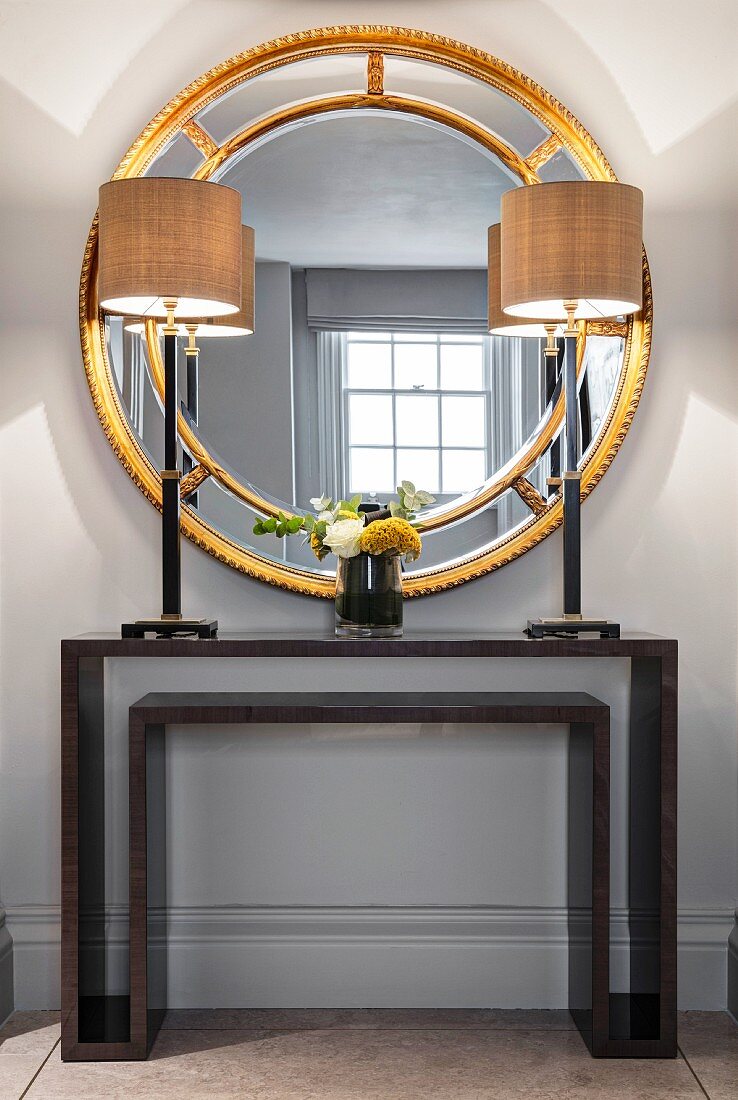 Round gilt-framed mirror in front of lamps on console table
