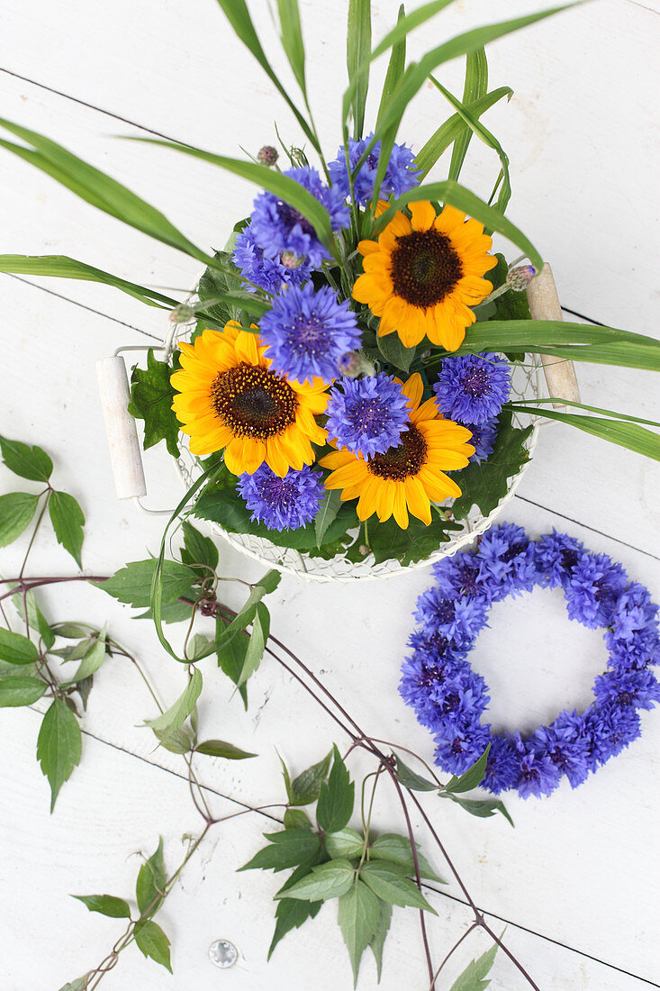 Sunflowers, cornflowers, blades of grass and clematis tendrils in wire basket next to wreath of cornflowers