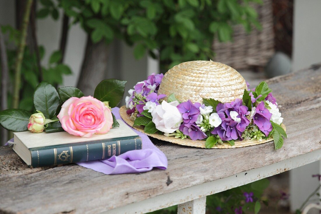 Strohhut mit Blumenkranz neben einem Buch mit Rose im Garten