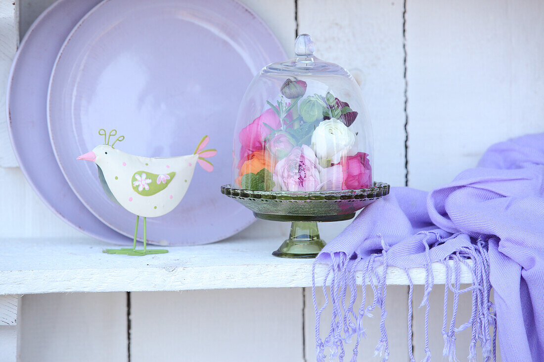 Colourful ranunculus under glass cover next to fringed lilac scarf and bird ornament