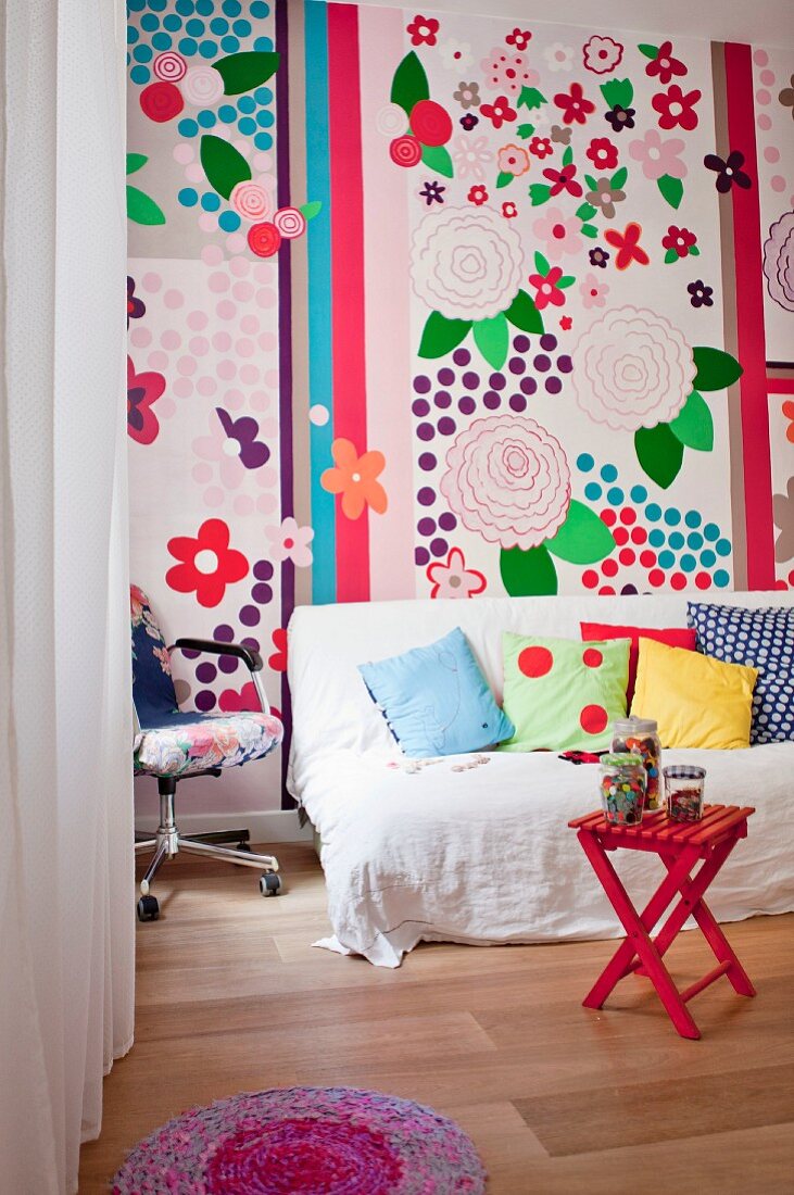 Couch mit weißem Überwurf und verschiedenen Kissen vor bunter Wanddekoration mit großformatigen floralen Motiven