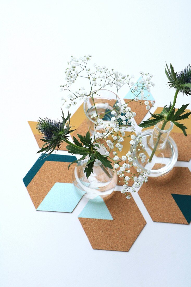 Vasen mit Distelblüten und Schleierkraut auf sechseckigen, bemalten DIY-Korkuntersetzern