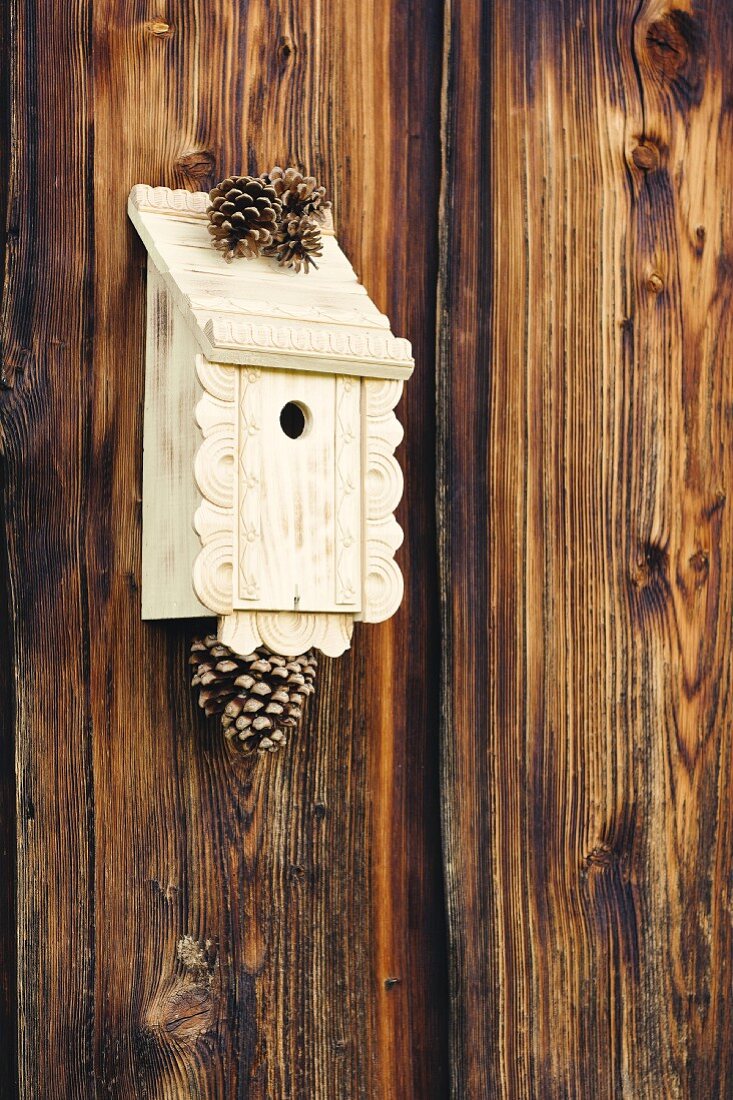Traditional bird nesting box decorated with ornamental trim and pine cones hung on board wall