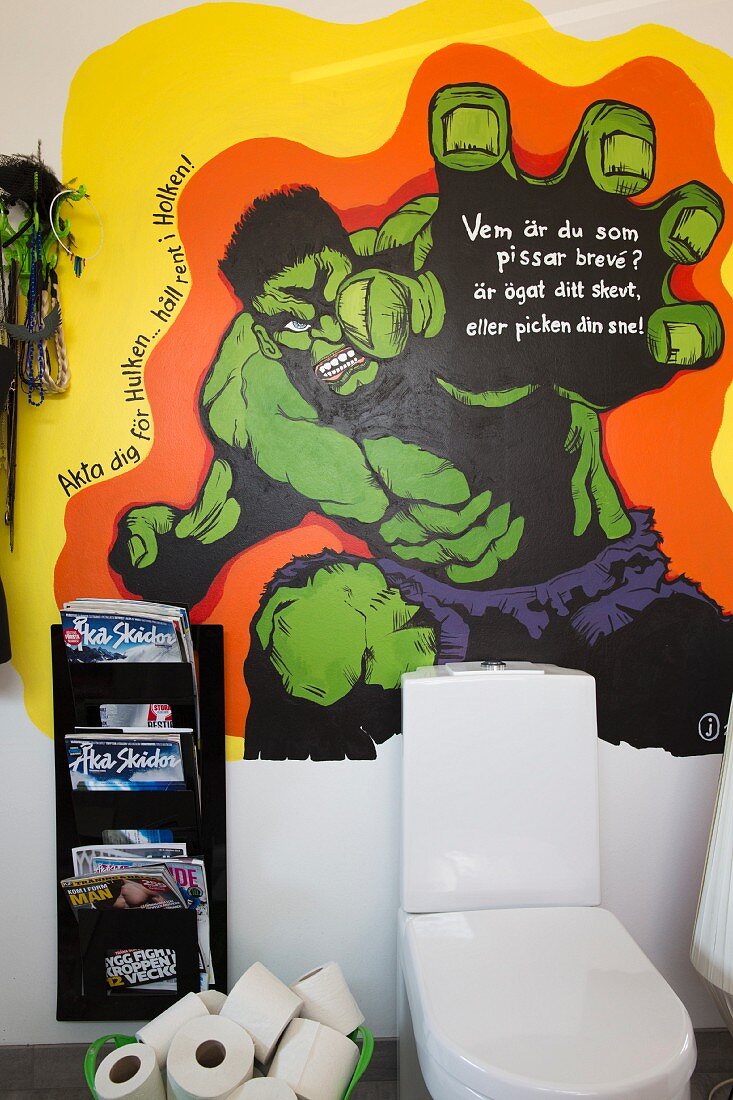 Toilette mit Spülkasten und Zeitschriftenhalter vor bemalter Wand mit Comicmotiv