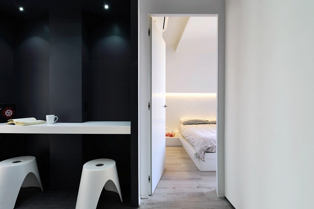 Massgefertigte weiße Theke an schwarzer Wand dazu weiße Hocker und offene Schlafzimmertür