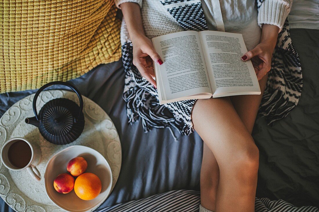 Frau liegt lesend auf Bett, daneben Tablett mit Tee und Obst