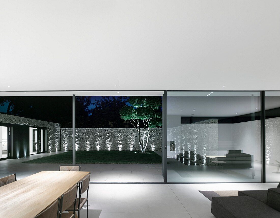 View from designer living area through open glass wall into courtyard with illuminated stone wall, terrace and lawn at twilight