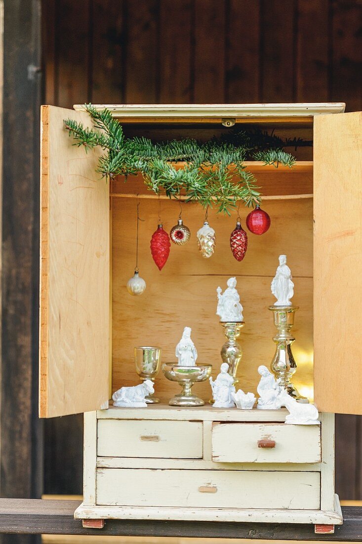 Modern nativity scene arrangement of white-painted nativity set figurines, old Christmas tree decorations, mercury silver candlesticks and green fir branches in vintage dolls'-house cabinet