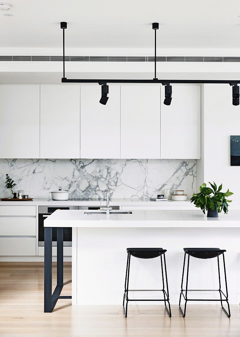 Schwarze Barhocker vor weisser Kücheninsel unter Lichtschiene mit schwarzen Strahlern in offener Designerküche mit Marmorspritzschutz