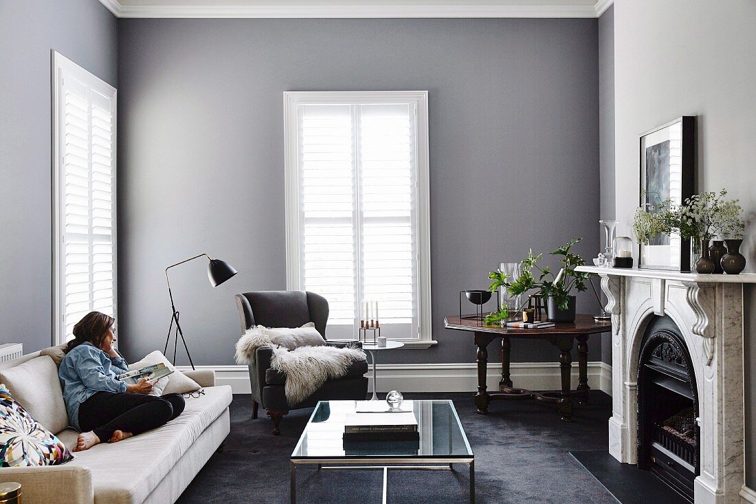 Elegantes, graues Wohnzimmer mit Frau auf Sofa vor modernem Glascouchtisch und Kamin