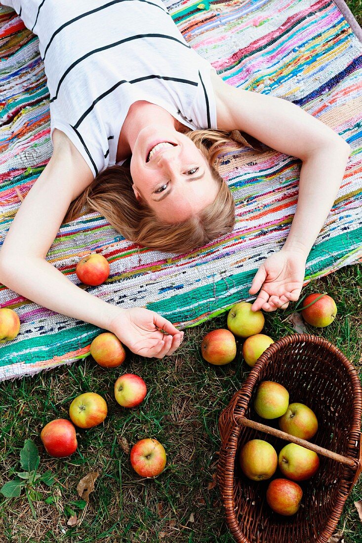 Frau auf einem Webteppich liegend, über ihrem Kopf frisch geerntete Äpfel