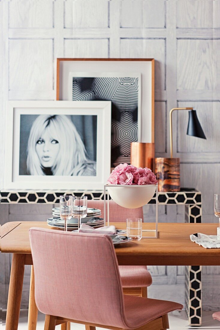 A bowl of pink flowers on a dining table in front of a black and white console table with framed pictures