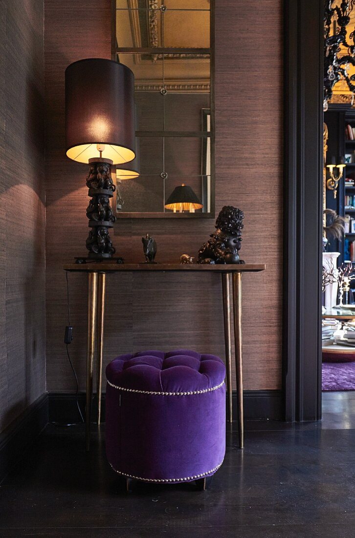 Violetter, eleganter Sitzpouf vor Wandtisch mit kunsthandwerklicher Tischleuchte und Tierfiguren