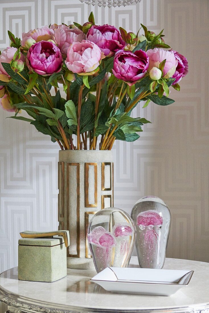 Vase of peonies and glass ornaments on side table in front of patterned wallpaper