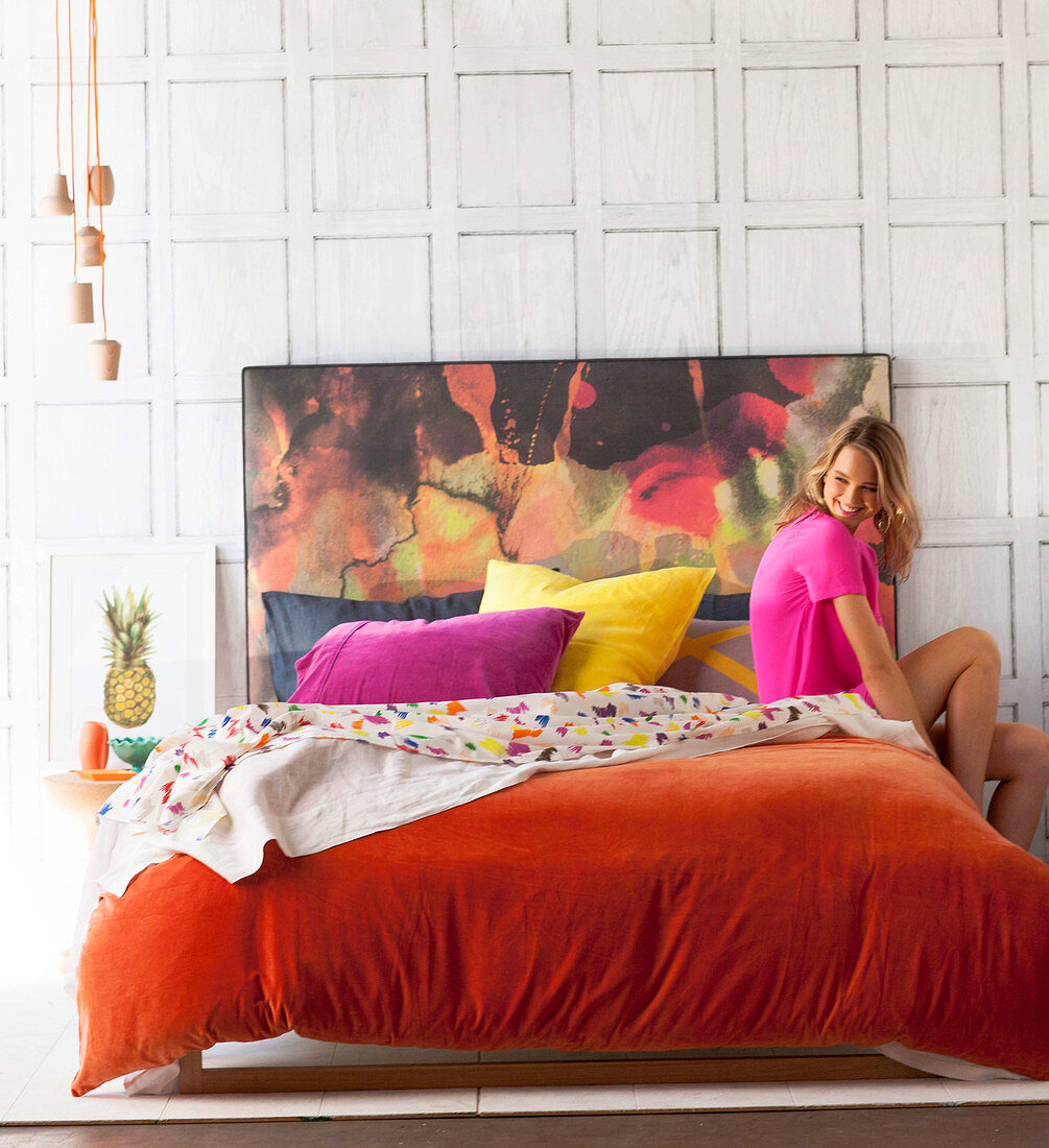 Junge Frau auf Doppelbett mit orangefarbenem Samtbezug und buntem Betthaupt