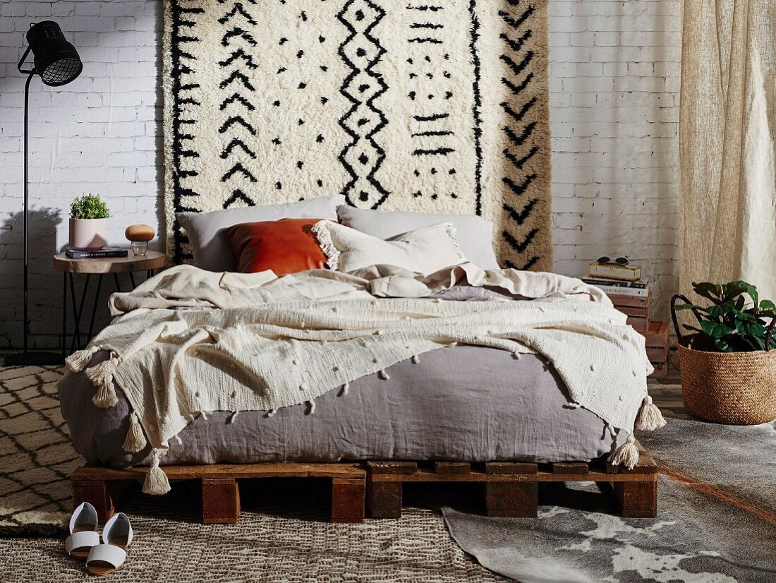 Doppelbett auf rustikalen Paletten vor weißer Ziegelwand mit Wandteppich