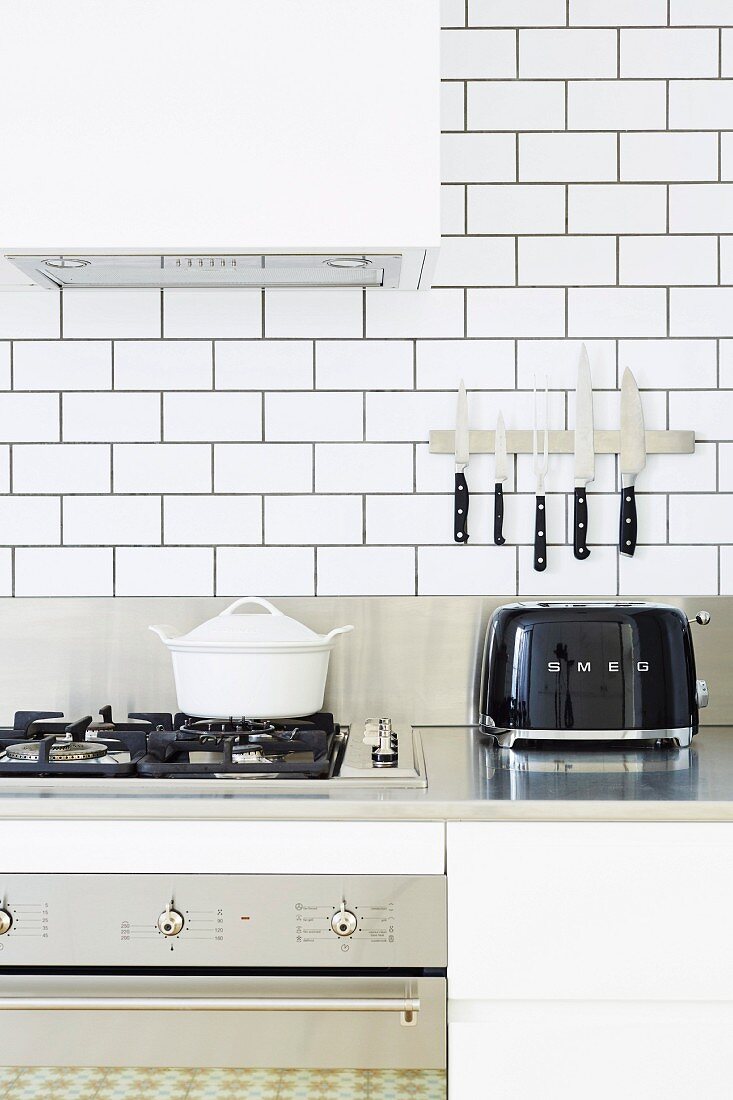 Weiß geflieste Küche mit Edelstahlarbeitsplatte, Gasherd und schwarzem Toaster unter Magnetschiene für Messer