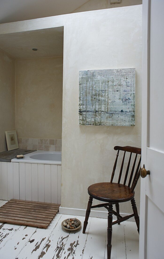 Blick auf Holzstuhl, im Hintergrund Badewanne teilweise sichtbar, schlichtes Bad mit Dielenboden, weiße abblätternde Farbe