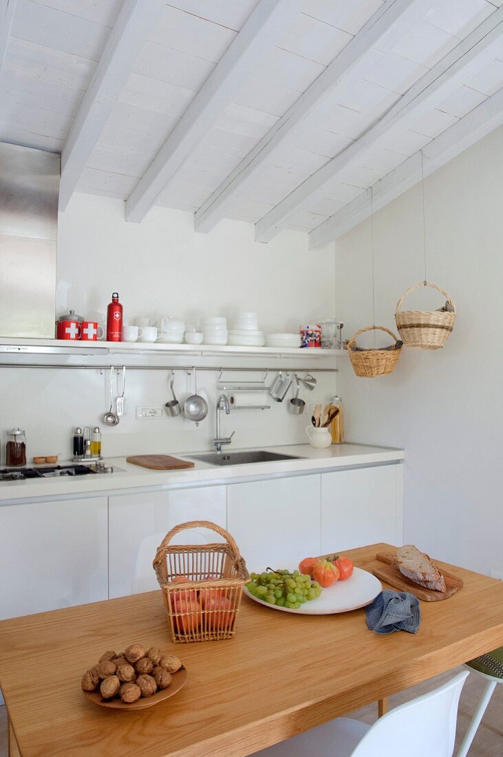 Walnuts and fresh fruit on wooden table in front of white kitchen counter below white wood-beamed ceiling