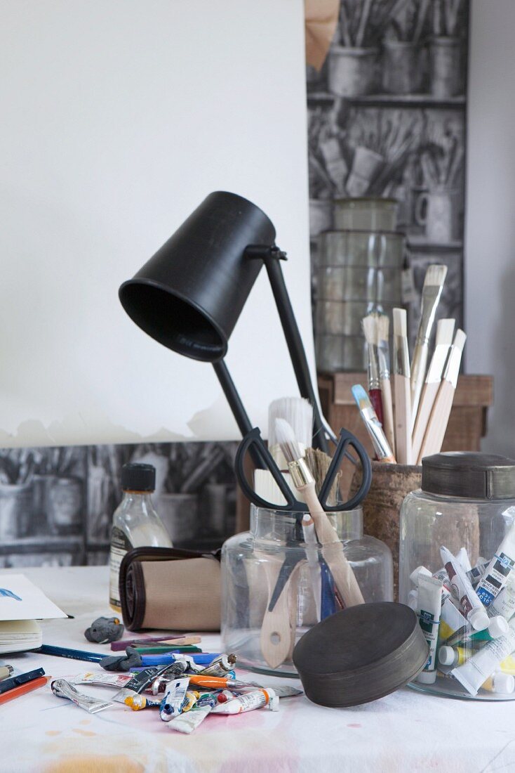 Verschiedene Aufbewahrungsgläser mit Künsterutensilien und Farbtuben auf Arbeitstisch mit schwarzer Tischleuchte