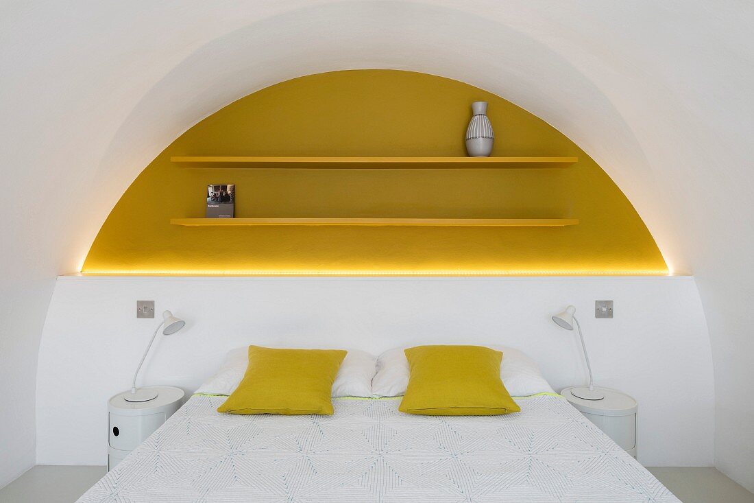 Doppelbett mit gelben Kissen vor halbhoher weißer Wand, darüber gelb lackierte Regalböden in hinterleuchteter gleichfarbiger Rundbogennische
