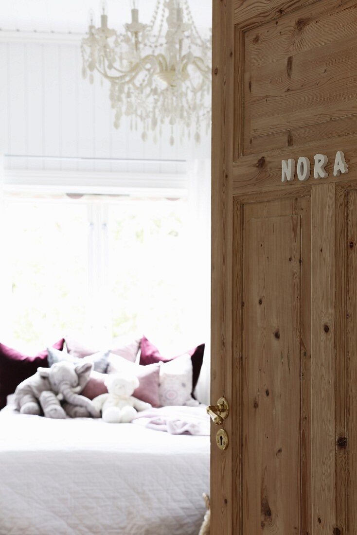 Offene Zimmertür mit Namen, Blick auf Bett mit Stofftieren und weißem Kronleuchter