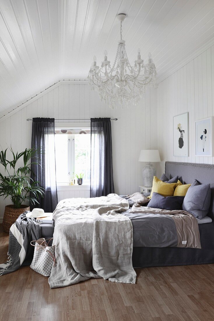 Blankets and cushions on box-spring bed with grey headboard in bedroom with white wood cladding