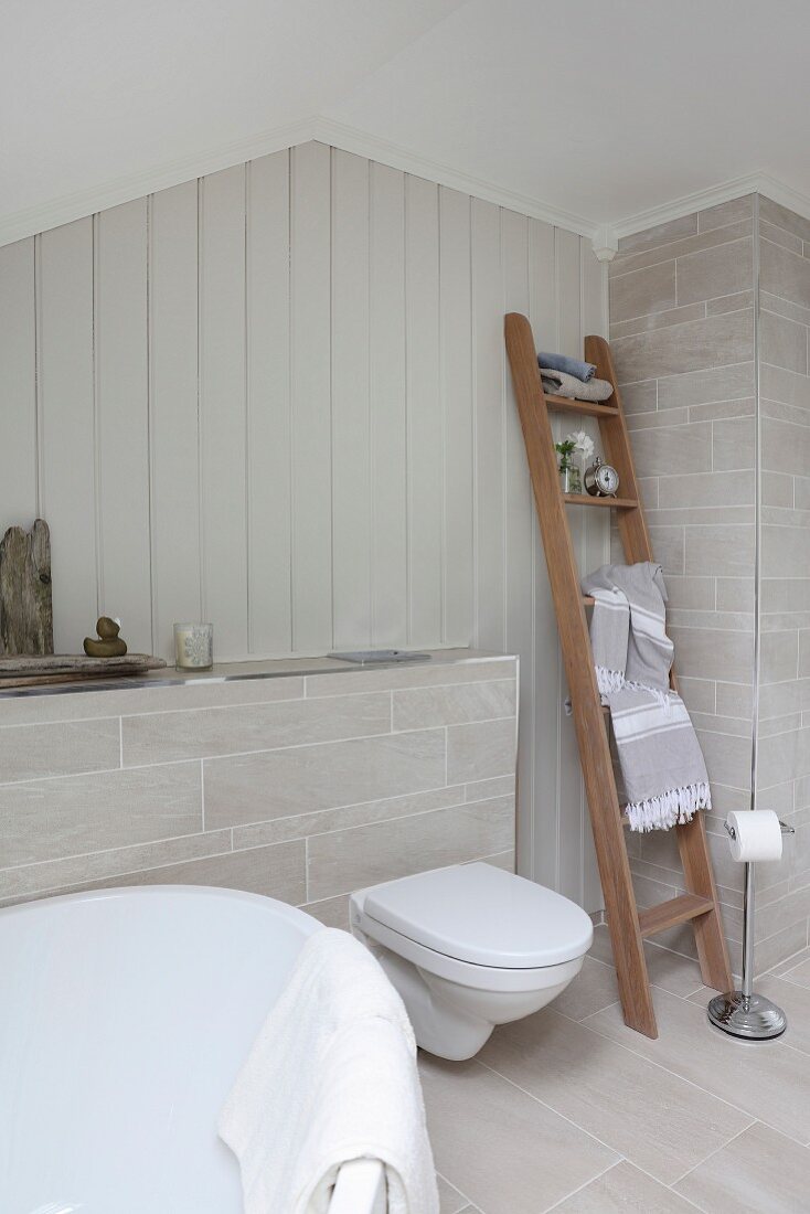 Bad mit naturfarbenen Fliesen an Vormauerung und Boden, Holzleiter mit Handtüchern neben Toilette