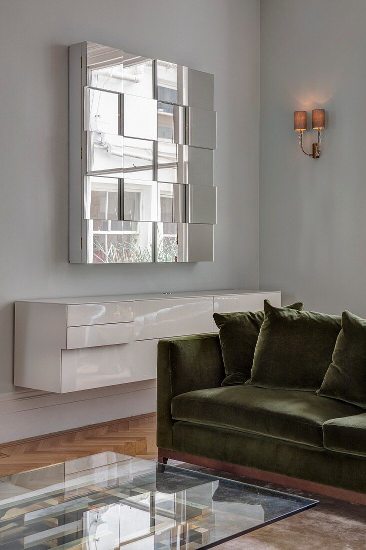 Glass coffee table, velvet sofa, white floating sideboard and mirrored wall-mounted cabinet in corner of living room