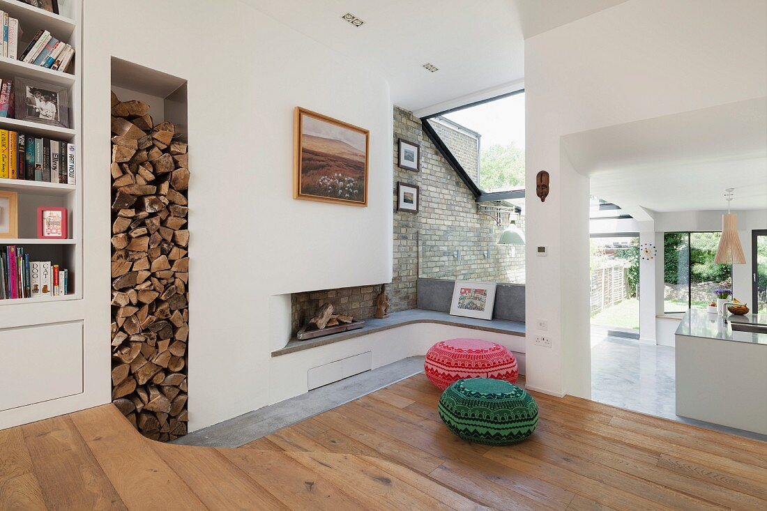 Moderner Wohnbereich mit Holzboden, vor Kaminecke farbige Sitzpoufs und Durchgang mit Blick in offene Küche