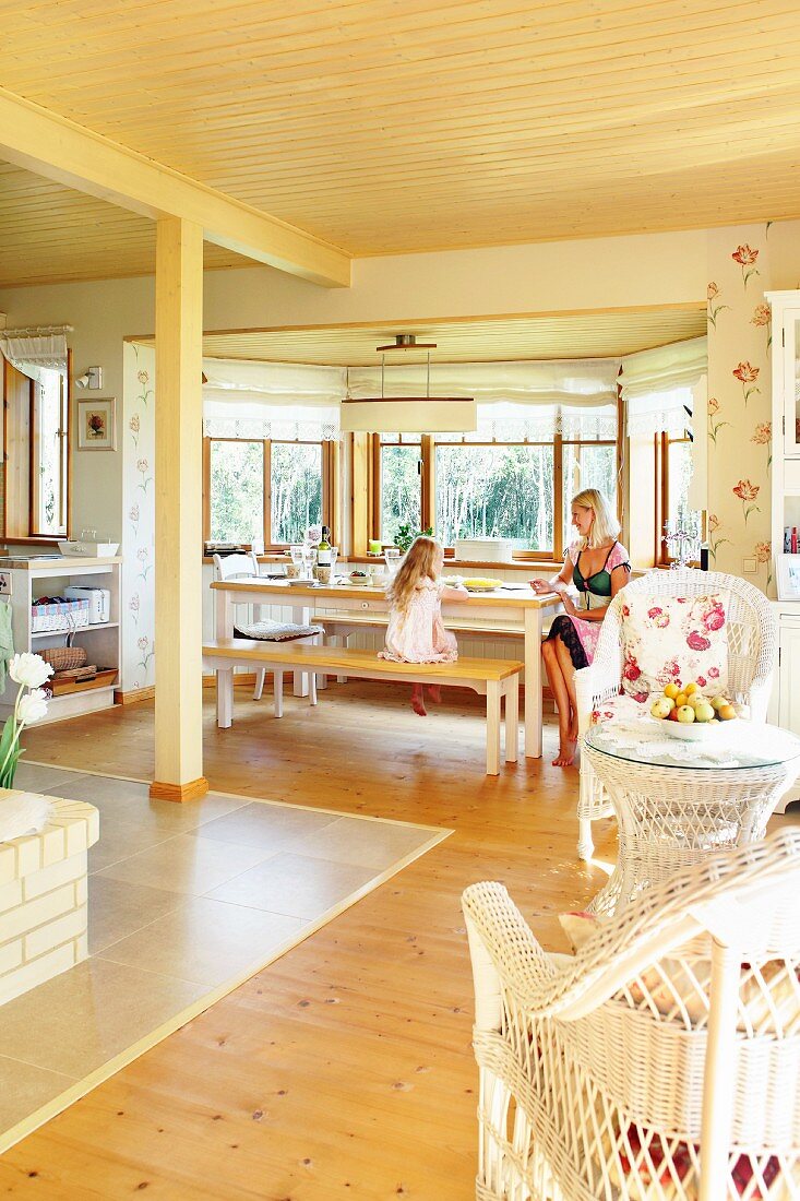 Offener Wohnraum mit heller Holzdecke, Korbmöbeln und Erker mit Mutter und Tochter am Essplatz