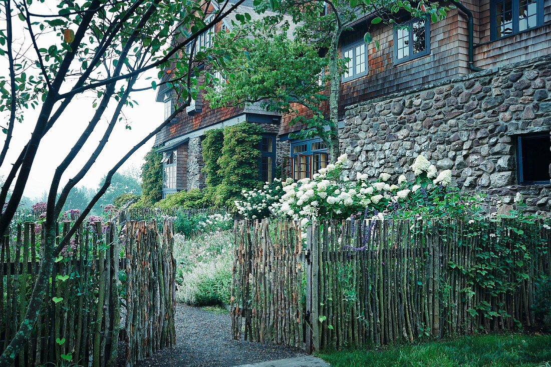 Knüppelzaun mit offenem Tor, weiße Fliederbüsche vor altem Landhaus teilweise mit Naturstein und Schindel Fassade