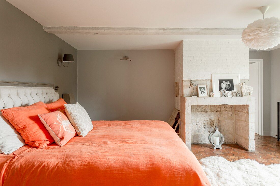 Double bed with orange bed linen in traditional bedroom with masonry, disused fireplace