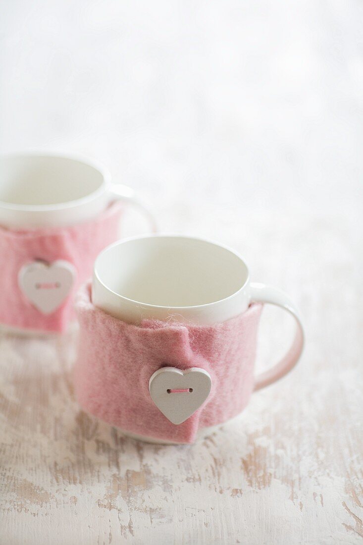weiße Tassen mit rosafarbenen DIY-Filzwärmern aufgenähten herzförmigen Knöpfen