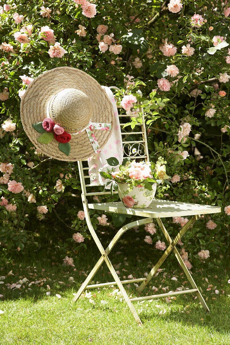 Flower arrangement and romantic straw hat on vintage garden chair in front of pink climbing rose