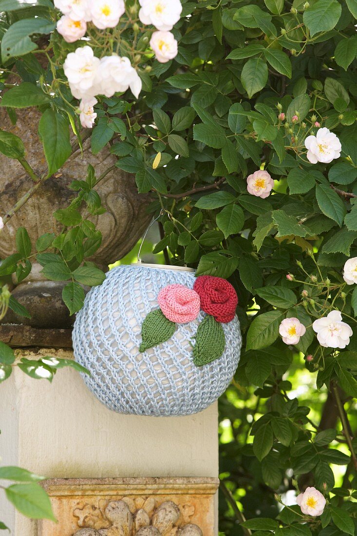 Romantic, knitted lantern decorated with knitted flowers hung below white climbing rose