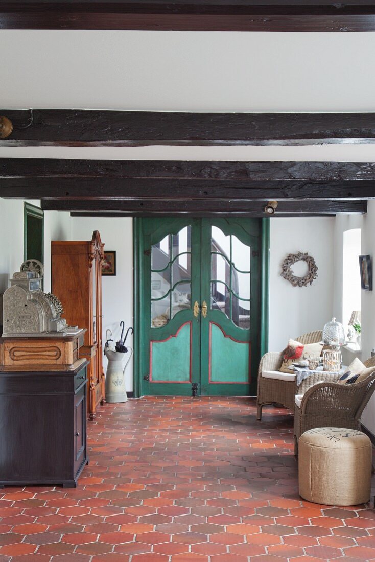 Green double doors, wicker armchairs, antique furniture and terracotta floor tiles in hallway of renovated farmhouse