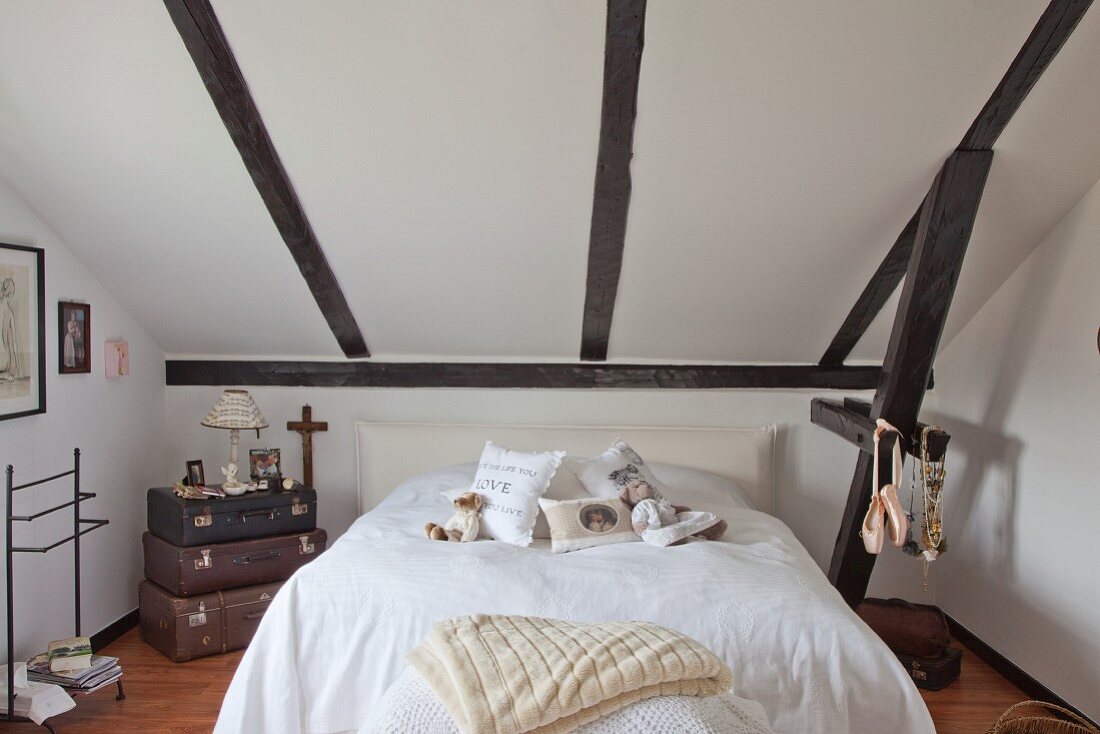 Teddy bear and scatter cushions on double bed next to stacked vintage suitcases below sloping ceiling in renovated farmhouse