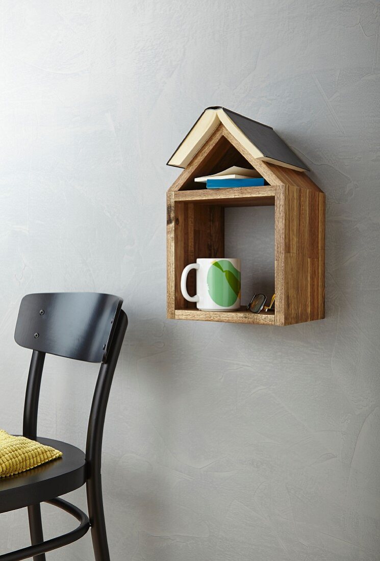 Books, cup and glasses on hand-made, wooden, house-shaped shelf next to chair