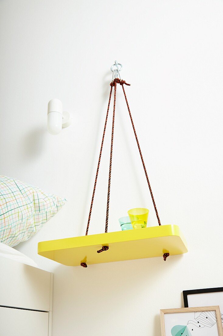 DIY shelf painted yellow hung on wall from ropes and used as bedside table