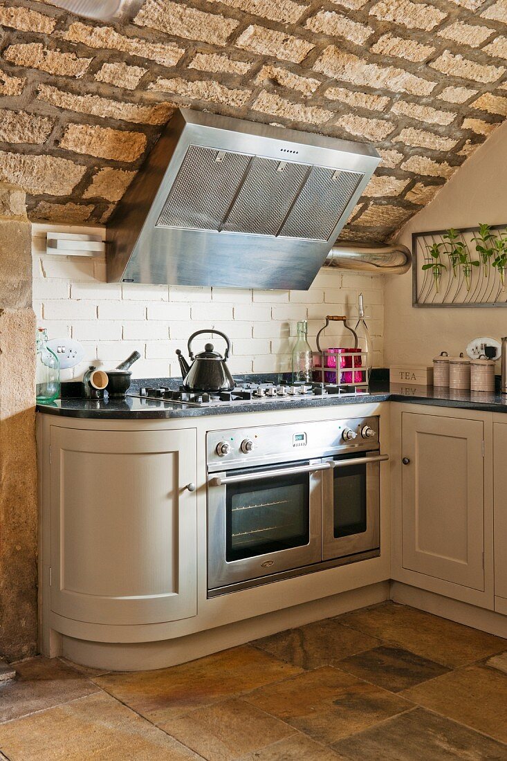 White country-house kitchen counter with rounded end cupboard and gas cooker under extractor hood on rustic, vaulted stone ceiling