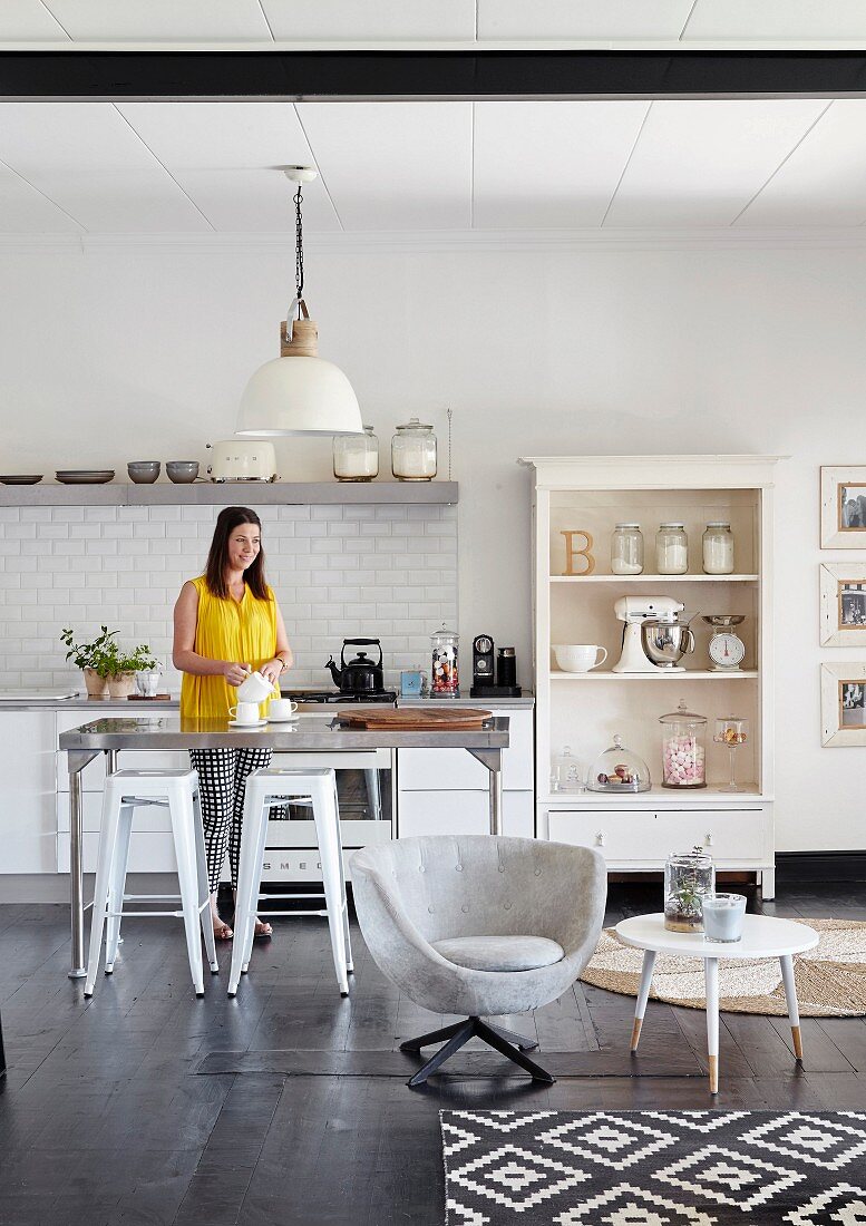 Woman next to metal table and bar stools in open-plan kitchen with open-fronted shelves