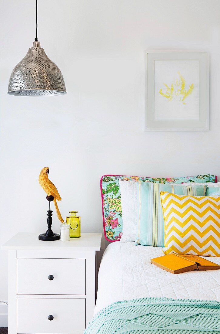 Bed with various pillows next to white bedside table with parrot figure
