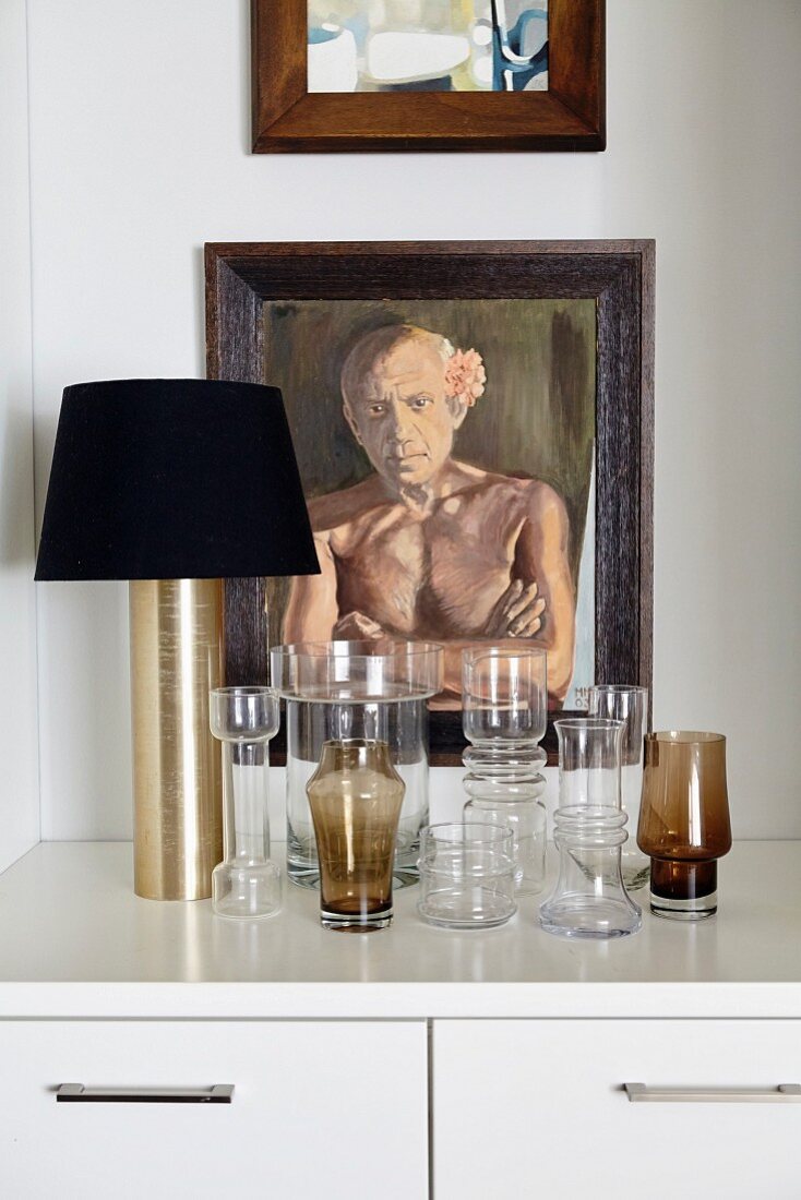 Collection of glasses and table lamp in front of framed portrait of man