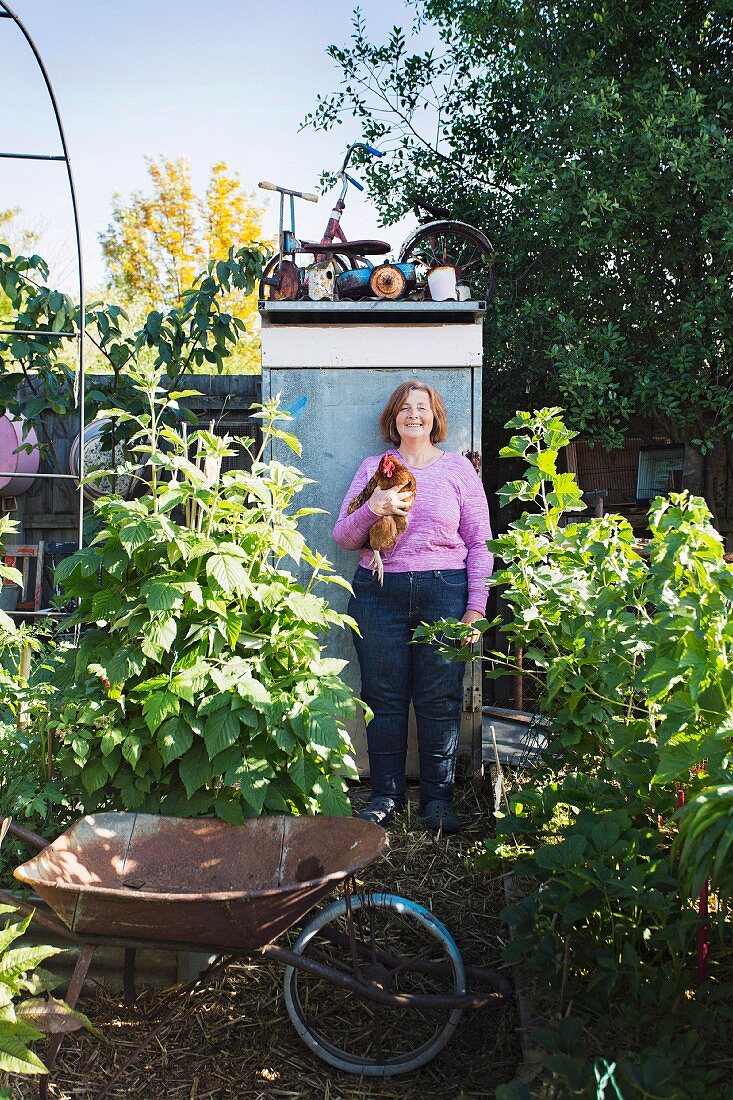 Woman with chicken in front of metal cupboard in garden with vintage wheelbarrow