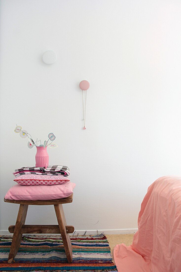 Rustikaler Holzhocker mit rosafarbenem Kissenstapel auf Flickenteppich in Schlafzimmer, Halskette an Wandhaken