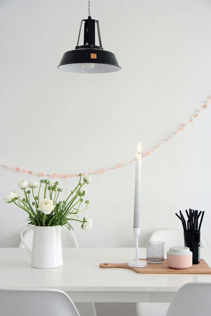 Ranunkelstrauss, Kerze und schwarze Trinkhalme auf weißem Esstisch vor rosa Girlande