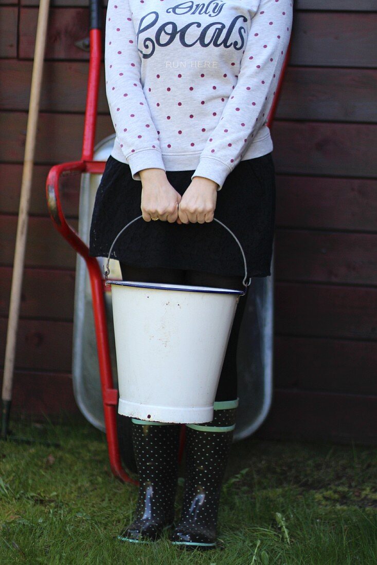 Woman wearing Wellington boots and holding enamel bucket stood outside garden shed